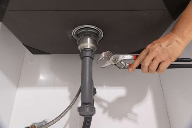 Main tenant une clé à molette pour serrer le raccord d'un siphon en PVC gris sous un évier, afin de réparer une fuite sous l'évier.