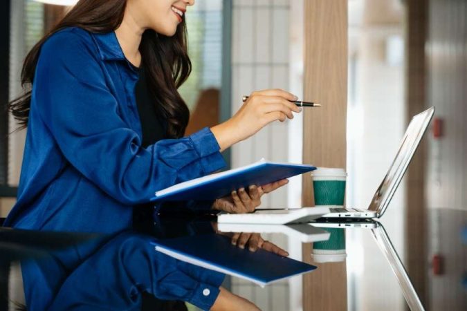Une professionnelle souriante en veste bleue examine un document devant son ordinateur portable.