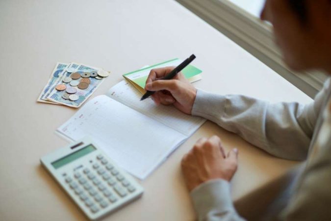 Une personne écrit dans un carnet de comptes, entourée d'une calculatrice et de monnaie japonaise, pour illustrer la méthode pour bien gérer son budget.
