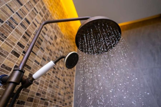 Une grande pomme de douche fixe au design rétro diffuse un large jet d'eau pétillant devant un mur de carrelage en mosaïque marron.